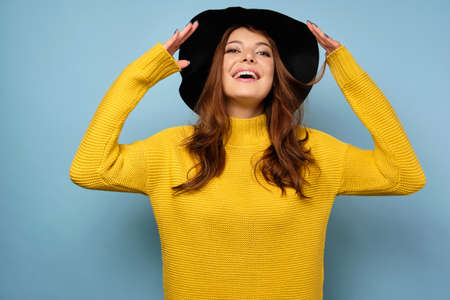 A brunette stands on a blue background in a yellow sweater and smiles with head thrown back and the edges of the black hat raisedの写真素材