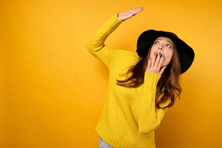 The brunette stands on a yellow background in a yellow sweater and black hat, looks with horror at...