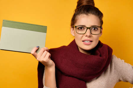 A brunette with glasses and a burgundy scarf, with a high bun, stands with a book in hand and looks displeasedly into the frame.の写真素材