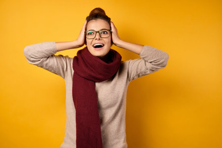 A brunette in a scarf and glasses with a high beam looks shocked at the top, clutching her hands to...