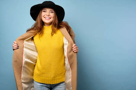 A brunette in a yellow sweater and black hat stands on a blue background, revealing a beige coat to the sides.の写真素材