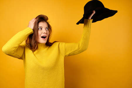 The brunette in a yellow sweater, opening her mouth in surprise, looks at the black hat raised in her hand whileの写真素材