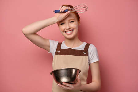 Cute red-haired girl with a high bun in a beige apron and a face in flour stands on a pink background, laughs with a bowl in handsの写真素材