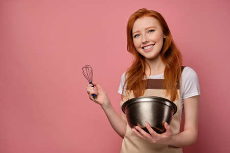 The red-haired girl in a beige apron and T-shirt stands on a pink background, holds a bowl and a whisk in hands, smiles broadlyの写真素材