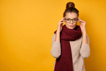 A beautiful brunette with a bun on head and glasses stands on a yellow background in a sweater and a scarf, looks at the cameraの写真素材