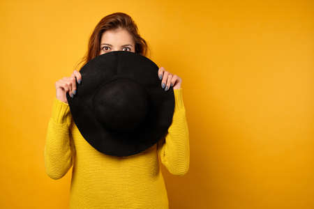 A beautiful brunette in a yellow sweater stands on a yellow background, covering her face to the eyes with a black hat.の写真素材
