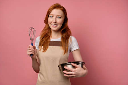 A red-haired girl in a beige apron and t-shirt is smiling on a pink background, holding a whisk and clutching a bowl to herselfの写真素材