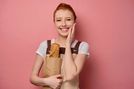 A cute redhead girl with a high bun in a apron and a face in flour stands on a pink background laughs, clutching a bag with a loafの写真素材