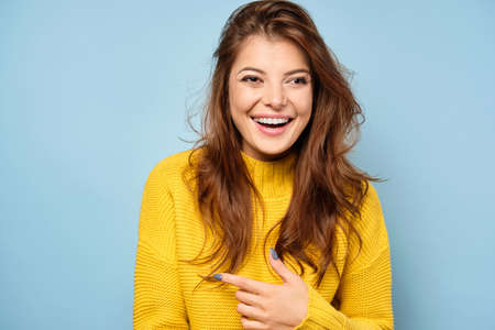 A beautiful brunette in a yellow sweater stands on a blue background, laughs and points a finger to the side.の写真素材