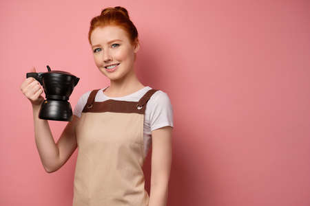 Cute redhead girl with a high bun in a apron stands on a pink background and smiles into the frame, holding a coffee pot in handの写真素材
