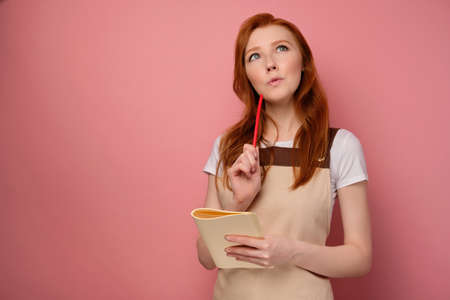 A beautiful red-haired girl in an apron holds a Notepad in her hand and looks up thoughtfully with a pencil to her lips.の写真素材
