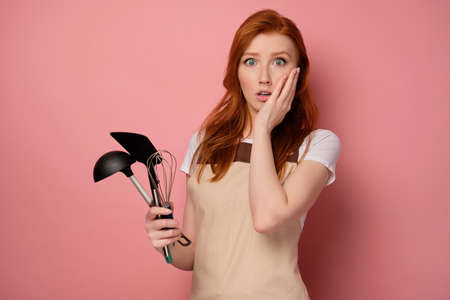 A red-haired girl in a apron on a pink background, holds kitchen appliances in hand and with surprise, presses hand to faceの写真素材