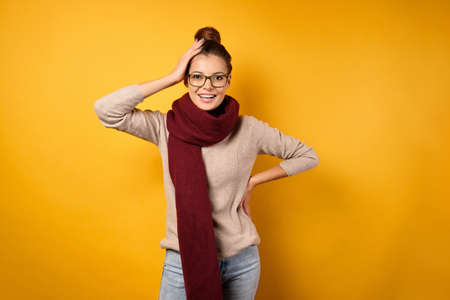 A beautiful brunette with a bunch on head and glasses stands in a sweater and a burgundy scarf, smiling, holds on to headの写真素材