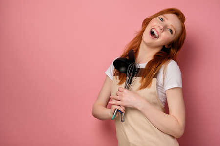 A red-haired girl in a apron stands on a pink background, joyfully opening mouth, clutching kitchen appliances to herselfの写真素材