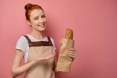 A cute red-haired girl with a high bun in a beige apron, with her face in flour and smiling, points to a packet with a loafの写真素材