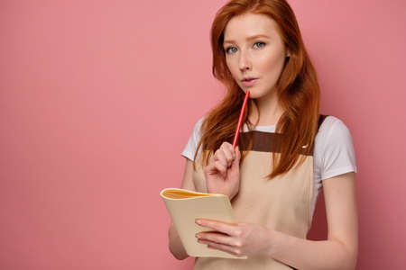 A red-haired girl in a apron stands on a pink background with a notebook and looks at the camera, applying a pencil to lipsの写真素材