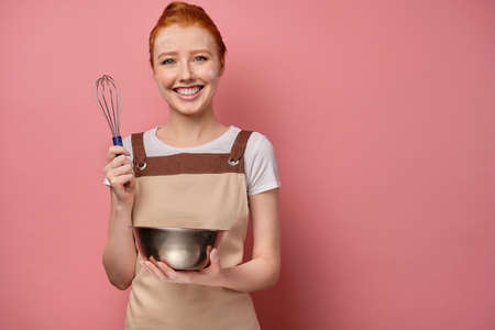 A cute red-haired girl with a high bun in a beige apron and a face in flour smiles broadly, holding a whisk and a bowl in handsの写真素材