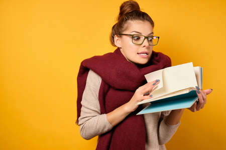 A brunette with a high bun and glasses stands on a yellow background in a sweater and scarf, flips through a notebook in horrorの写真素材