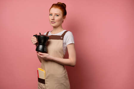 A cute red-haired girl with a high bun in a beige apron stands on a pink background, with a coffee pot in her handsの写真素材