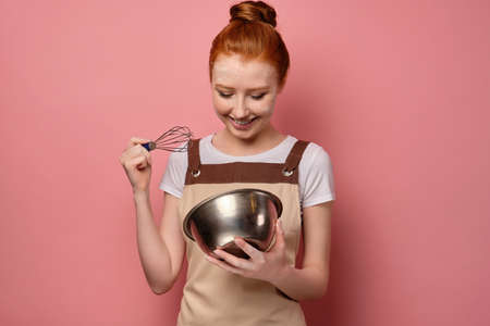 Cute red-haired girl with a high bun in a beige apron and a face in flour smiles broadly, looking into a bowlの写真素材