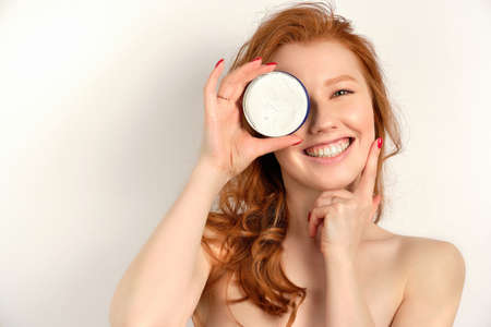 A red-haired curly-haired girl in a pink towel stands on a white background, covering eye with a jar of cream and smiling widelyの写真素材