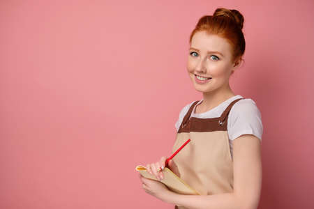 A red-haired girl in an apron with collected hair stands in a half-coat on a pink background with a notebook and pencil and smilesの写真素材