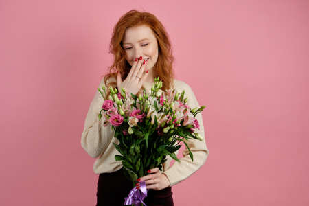 A red-haired curly girl in a white sweater stands on a pink background holding a bouquet in hand and smiling, hiding behind palmの写真素材