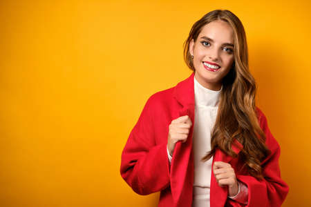 A beautiful girl with clean skin and red lipstick stands on a yellow background and corrects the lacquers of a red coatの写真素材