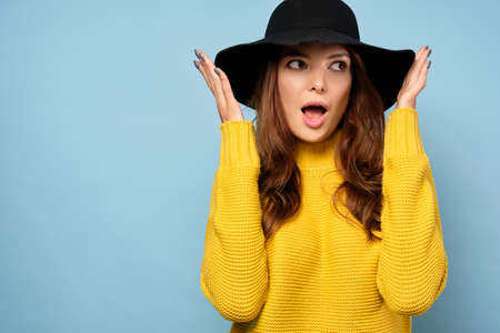 A brunette in a yellow sweater stands on a blue background, looks in astonishment to the side and holds the hat with her handsの写真素材
