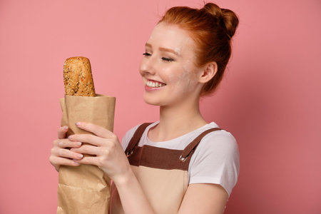 Redhead girl with gathered hair, in an apron, stands on a pink background, smiling and looks at a...