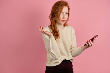 A red-haired girl with red lipstick on a pink background with a phone in hand and is looking upward pursed lips in displeasureの写真素材