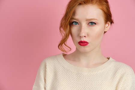 A beautiful red-haired girl with red lips in a white sweater stands on a pink background and squeezes lips looking at the cameraの写真素材
