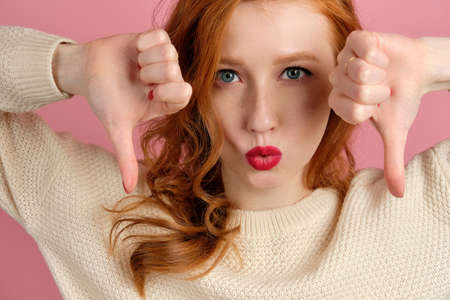 A close shot of the face of a red-haired girl with red manicure and lipstick looking at the frame and lowering the thumbsの写真素材