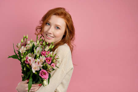 Beautiful red-haired girl hugs a bouquet with flowers on a pink background and gently smiles at the cameraの写真素材