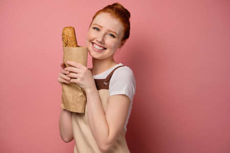 Beautiful redhead girl with gathered hair in an apron, with flour on her face, smiles broadly holding a bag of bread in her handsの写真素材