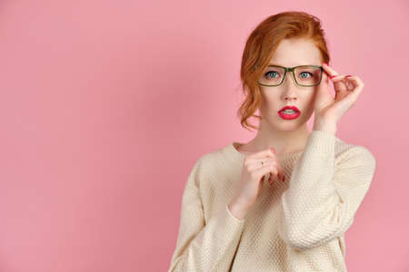 Beautiful redhead girl with red lipstick and manicure stands on a pink background and adjusting his glasses, looking at the cameraの写真素材
