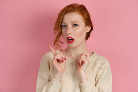 Beautiful red-haired girl with red lips in a white sweater stands on a pink background and points a finger to the sideの写真素材