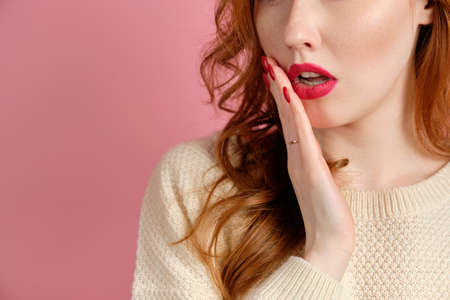 A close shot of the face of a red-haired girl with red manicure and lipstick holding her hand against parted lips, focus on the lips. Horizontal photoの写真素材