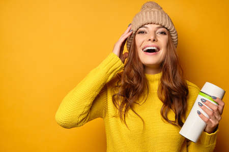 Beautiful brunette see clean skin in a yellow sweater and hat stands on a yellow background laughing joyfully holds a thermomugの写真素材