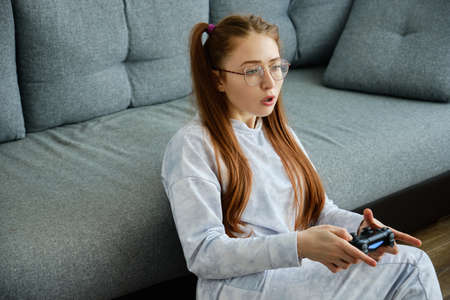A red-haired girl with glasses, with long tails, sits on the floor in a half-turn with a game console and looks forwardの写真素材