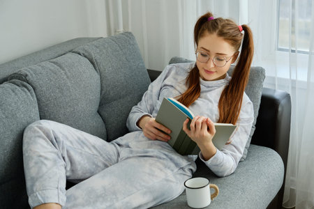 A red-haired girl with glasses, with long tails in pajamas lies on the sofa with a mug and reads a...