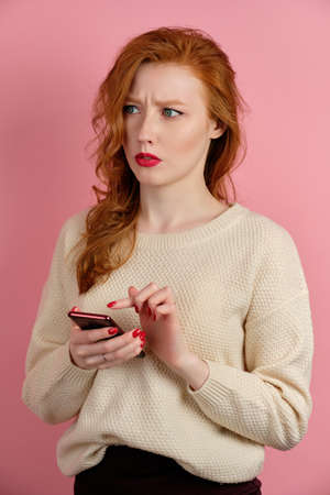 A red-haired girl with a phone in her hands is half a turn on a pink background and looks to the sideの写真素材