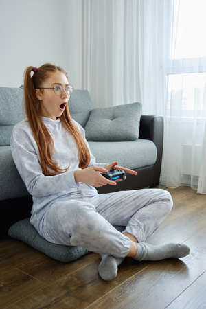 A red-haired girl with glasses with long tails, sits on the floor with a game console and opens mouth in shock, looking forwardの写真素材