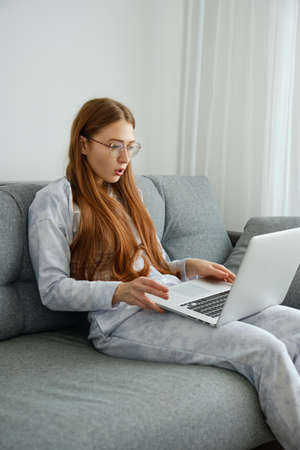 A red-haired girl with glasses, with long hair in pajamas sits on the sofa in a half-turn and looks at the laptopの写真素材