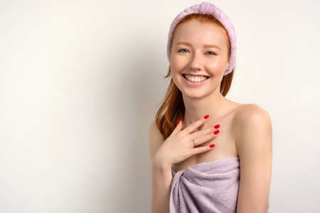 The red-haired girl with clean skin in a towel stands on a white background and smiles, holding her hand to her chestの写真素材