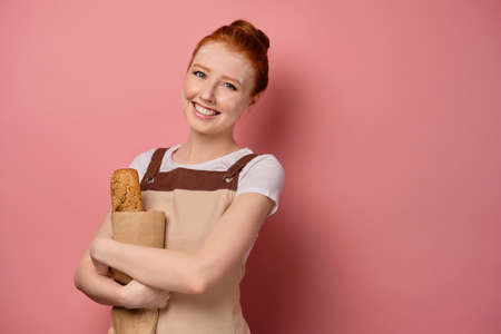 Red-haired girl with gathered hair, in an apron, is standing in an embrace with a bag of bread on a pink backgroundの写真素材