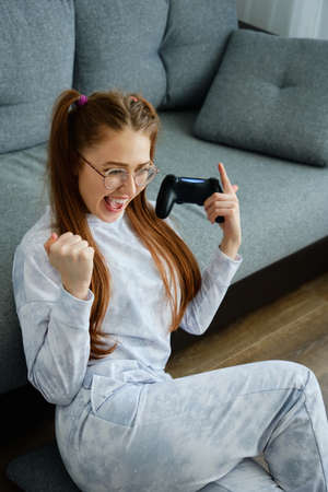 A red-haired girl with glasses, with long tails, sits on the floor joyfully raising her hands to her chest with a game consoleの写真素材