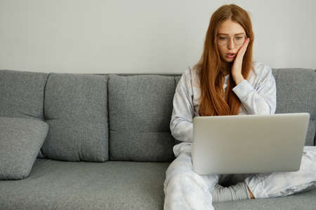 A red-haired girl with glasses, with long hair isits on the couch in a half-turn and looks intently at the laptop, opening mouthの写真素材