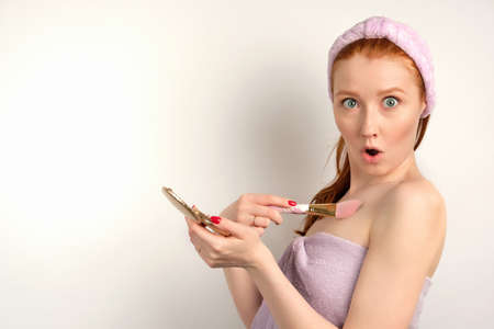 The redhead girl with clean skin in a towel stands with a small mirror in surprise looks into the frame, pressing a brush to chestの写真素材