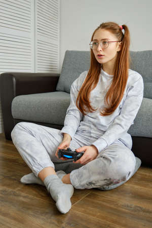 A red-haired girl with glasses, with long tails, sits on the floor in a half-turn with a game console and looks forwardの写真素材